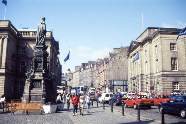 Gothic statue on Edinburgh High Street, cars parked beside stone buildings, pedestrians walking