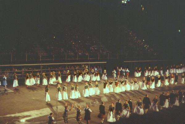 Dancers in white skirts and pipers march across a floodlit arena