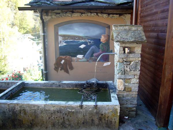 Old stone watering trough with flowing water, painted mural on wall behind