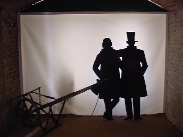 Two black silhouettes, one in top hat, beside an old wooden cart