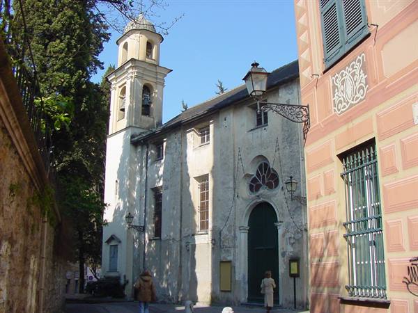 Church with bell tower among pastel houses, wrought-iron lamp, people walking in sunlight