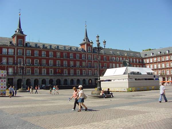 Tourists walk across Madrid's Plaza Mayor, red buildings and blue sky