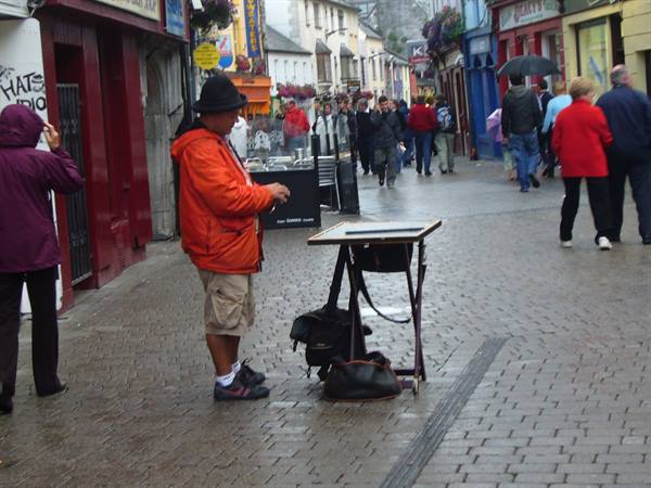 Busker in orange jacket and hat beside a small table on a cobbled street