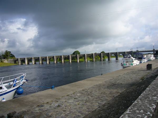 Boats moored beside a long stone arch bridge, cloudy sky over the river