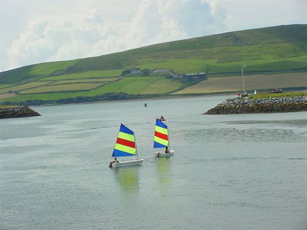 Two small sailboats with colorful sails glide past green hills