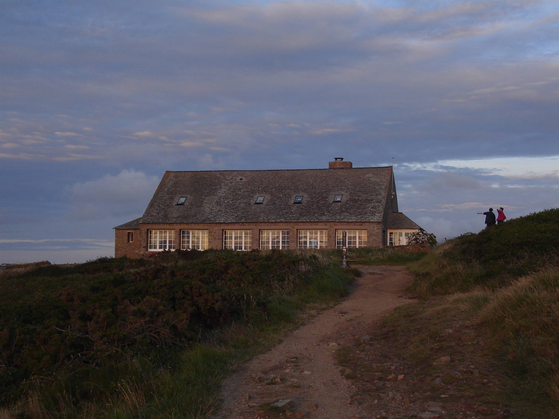Dirt path leading to a stone house with lit windows, two people walking at dusk