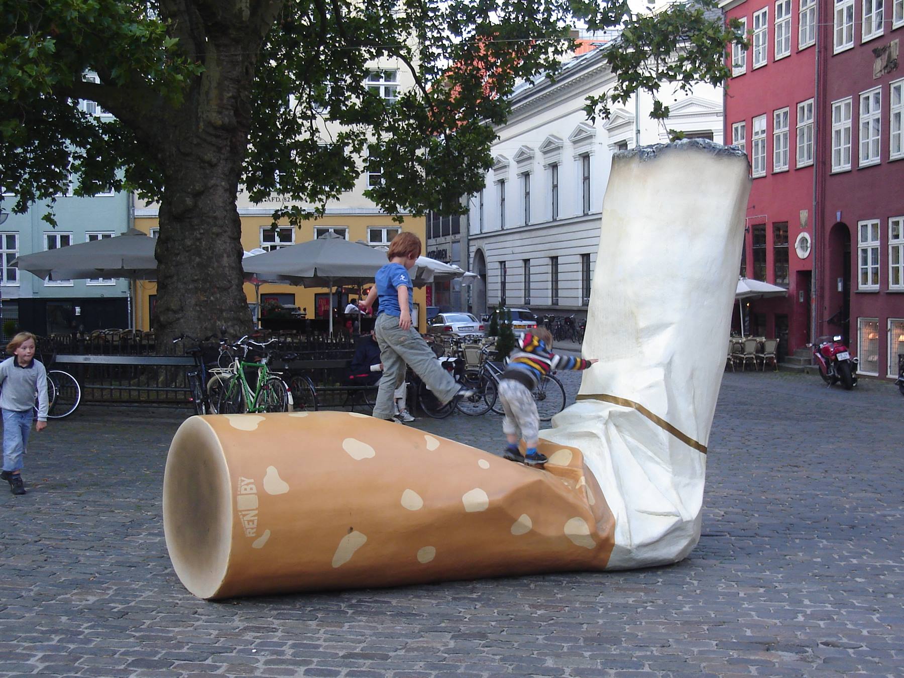 Children climbing a giant cigarette sculpture in a cobblestone town square