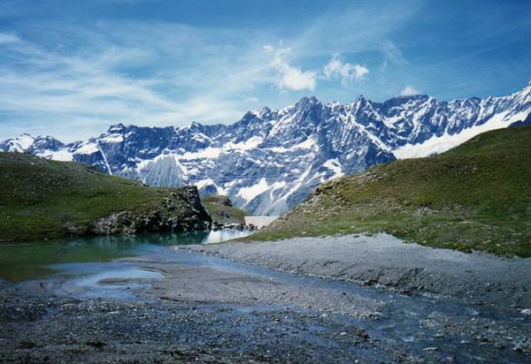 Milky alpine lake with grassy scree slopes and snow-capped peaks under blue sky