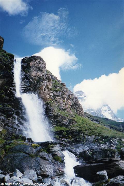 Foamy waterfall cascading over rocky cliffs, green meadow, blue sky with clouds