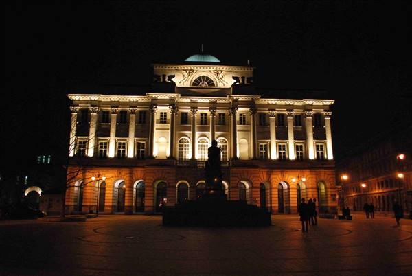 Neoclassical palace lit at night with equestrian statue in square, pedestrians