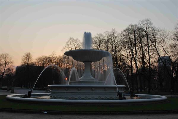 Two-tier stone fountain spraying water among bare trees, orange sky