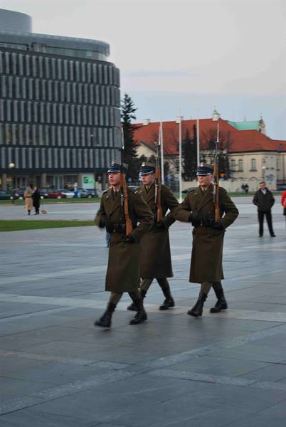 Three soldiers in green uniforms march with rifles across a paved square in Warsaw