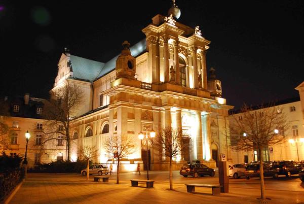 Baroque church lit up at night in Warsaw, benches and cars in foreground