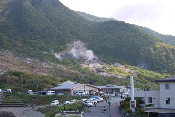 Volcanic steam rises from green mountain, buildings and visitors below, Owakudani