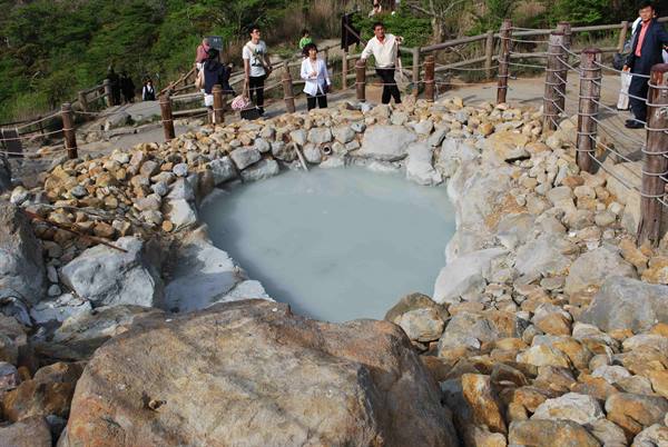 Tourists beside a milky hot spring pool surrounded by rocks and wooden fence