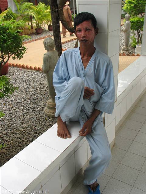 Man in light blue robe sitting on a tiled ledge, Buddha statues and plants around