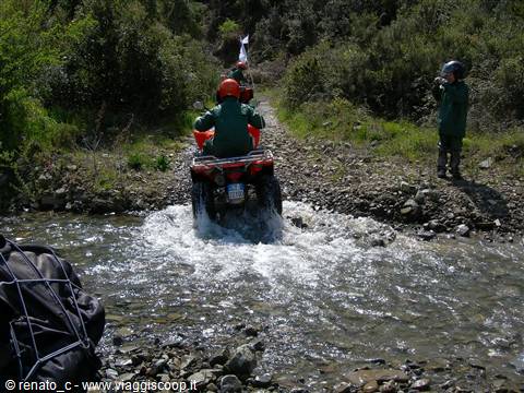 Red quad bike crosses a rocky stream among bushes, person in green watching