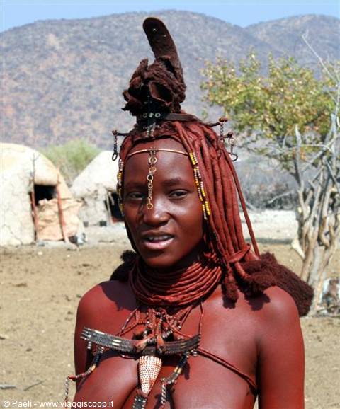 Himba woman with ochre braid and shell necklaces in front of huts