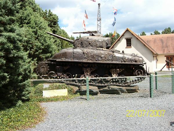 Rusty Sherman tank displayed behind a fence, with flags and a small house behind