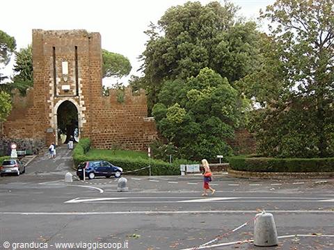 Woman crosses wet asphalt in front of Tuscania's tufa stone Porta Romana gate
