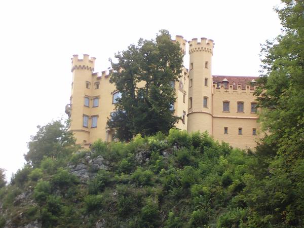 Yellow castle with crenellated towers above green rock, soft daylight