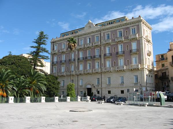 Pink palace with balconies across a stone-paved square, palms and parked cars
