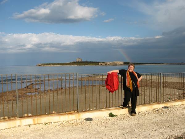 Woman with red jacket leaning on the railing before Isola delle Correnti, rainbow over the sea
