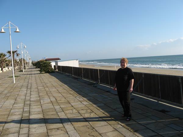 Woman in black on paved seaside promenade, lampposts and sandy beach in sunlight