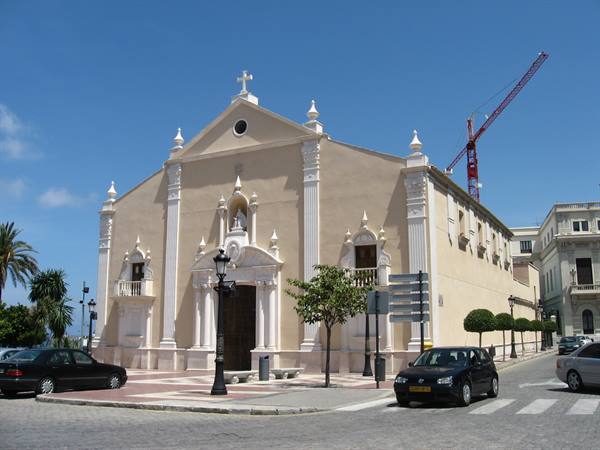 White and ochre baroque church, parked cars, palm trees and crane under blue sky