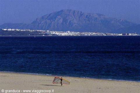 Windsurfer carrying his sail on a sandy beach facing deep blue sea and a mountain