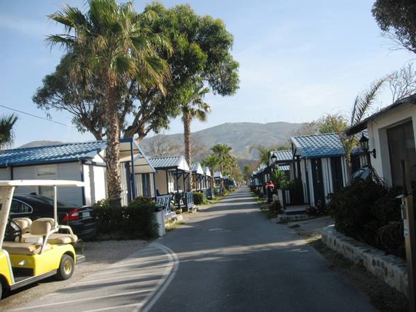 Bungalows with blue roofs along a paved lane, yellow golf cart, palms and hills