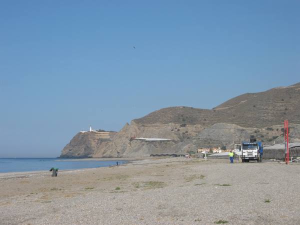 Truck on pebble beach, man in yellow vest, lighthouse on arid cliff