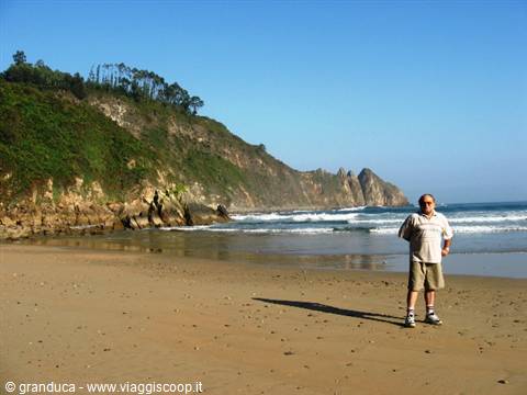Man in light shirt standing on sand, green cliffs and waves behind him