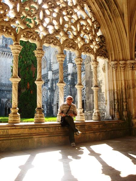 Man seated beneath the carved royal cloister of Batalha Monastery, sunlit stone columns
