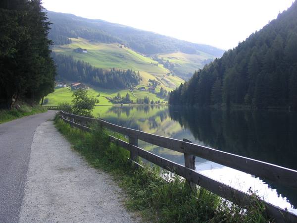 Gravel road along an alpine lake, wooden fence, forests and green meadows