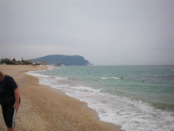 Woman walks along sandy pebbly beach, gentle waves and green headland under grey sky