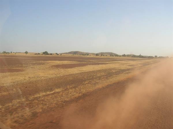 Red dust rising from a dirt road across dry savanna, distant hills under clear sky