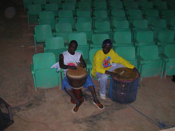 Two drummers play djembe and bass drum among green plastic seats