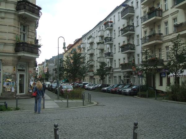 Blonde woman crosses a cobbled street between apartment buildings and parked cars