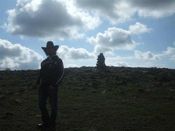 Man in hat backlit on grassy hill with stacked stone cairn
