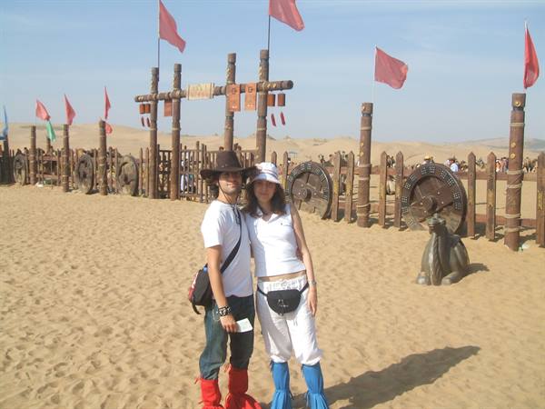 Couple in colored gaiters on sand before wooden gates with red flags, Kubuqi desert