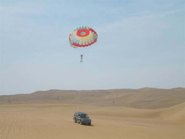 Tourist parasailing under a colorful canopy towed by an SUV over sand dunes