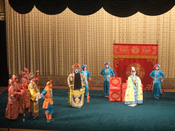 Actors in traditional Chinese costumes perform on a stage with embroidered red fabrics
