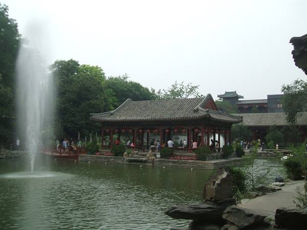 Wooden pavilion with grey tiled roof on lake, fountain, visitors under traditional Chinese roofs