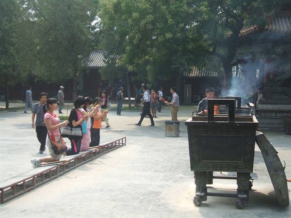Women kneel praying in a temple courtyard, smoking bronze incense burner nearby