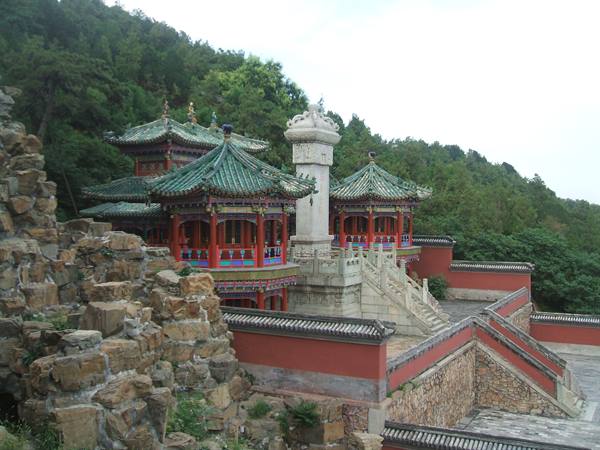 Pavilions with green roofs on red walls near white stone stupa, forested hillside