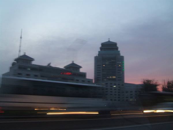 Illuminated tower behind traditional-roofed buildings, lights streaked from moving car window
