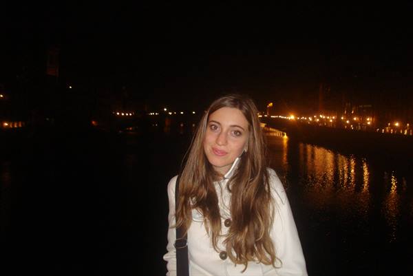 Young woman in white coat beside a river lit by night lamps