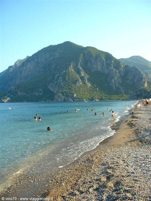 Swimmers in turquoise water on a pebble beach beneath green mountains