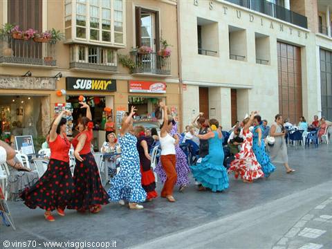 Women in floral dresses dance flamenco in a square outside cafes